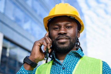 Afrikalı etnik köken mühendisi, rastalı ve sakallı, sarı güvenlik kaskı ve yeleği takan, telefonda konuşan, kameraya, fotokopi alanına, sanayiye ve insanlara bakan,.
