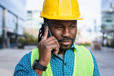 Afrikalı etnik köken mühendisi rastalı ve sakallı, sarı güvenlik kaskı ve yeleği takan, açık havada telefonla konuşan, mekanı, endüstriyi ve insanları taklit eden,.