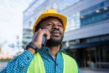 Afrikalı etnik köken mühendisi rastalı ve sakallı, sarı güvenlik kaskı ve yeleği takan, açık havada telefonla konuşan, mekanı, endüstriyi ve insanları taklit eden,.