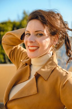 Günbatımında terasta durup mutlu bir şekilde gülümseyen ve kameraya bakan kırmızı dudaklı, kahverengi ceketli zarif Fransız kadının dikey portresi.