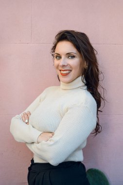 Beyaz ırktan beyaz bir kadının dikey portresi. Evinde durmuş gülümsüyor ve arka planda pembe duvara yaslanmış bir şekilde kameraya bakıyor.