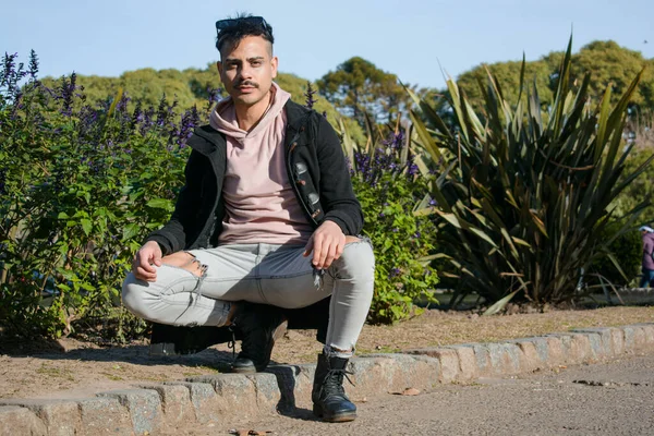 young fashionable homosexual latino man with mustache and short hair, wears pink sweater and black jacket, crouching posing looking at camera in park at sunset, lifestyle, copy space.