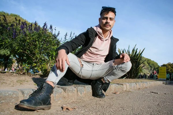 Arjantin kökenli yakışıklı homoseksüel Latin erkek portresi, bıyıklı ve kısa saçlı, açık bacaklı, halka açık parkta çalılar ve arka planda gökyüzü ile oturuyor, boşluğu kopyalıyor..