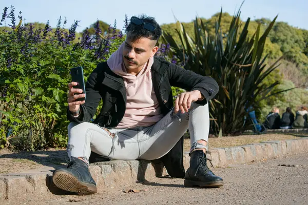young attractive latin fashionable man of argentinian ethnicity, sitting in park using phone, people and technology concept, copy space.