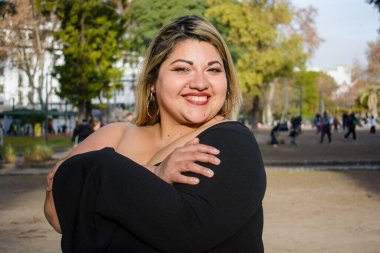 Arjantinli genç, iri yarı, Latin bir kadın gün batımında gülümseyen bir gülümsemeyle parkta kendine sarılıyor. insanlar kavramı, kopyalama alanı.