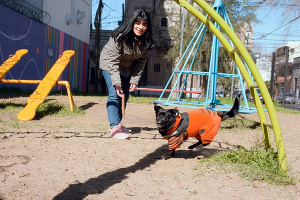 young black haired venezuelan latina woman, wearing jacket and jean pants, chasing her dog in park to grab her, playing with her pet on sunny day on weekend