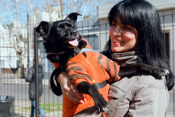 portrait of venezuelan latin young woman, black hair and winter clothes, happy smiling standing in park carrying her dog in her arms on sunny day, lifestyle and pet concept, copy space