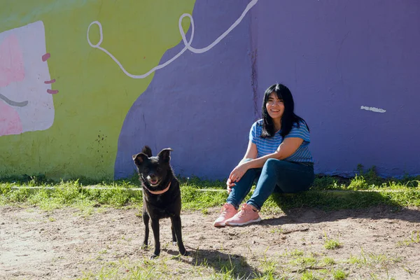 young latin venezuelan woman, in blue clothes, happy with her black dog in park at sunset, in buenos aires, smiling and looking at camera, pets and people concept, copy space.
