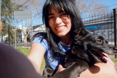 Gün batımında parkta köpeğiyle oturup kameraya gülümseyen genç Latin kadının selfie portresi..