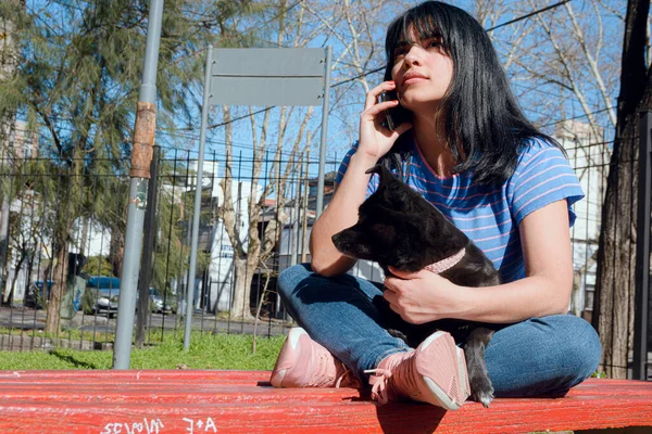 young Latin Venezuelan woman with black hair, dressed in blue, sitting in park with her attentive dog talking on phone at sunset, technology concept, copy space.
