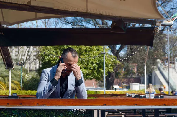 Zarif takım elbiseli yetişkin beyaz iş adamı, öğle vakti restoranın dışında oturup telefonla ve düşünerek endişeleniyor..