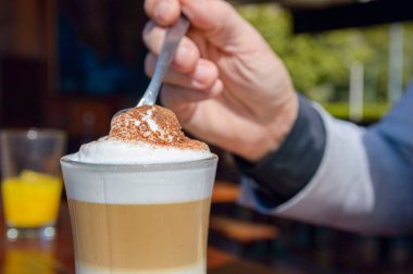 Kahvaltı sırasında restoranın dışında kaşık kaşık kapuçino köpüğü karıştıran beyaz erkek eli.