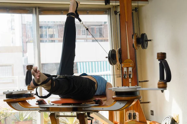 side view of woman lying on her back training her balance and power on exercise machine at home using pulleys by raising and lowering her legs