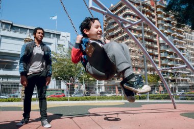 Genç afro Latin babası, mutlu bir şekilde gülüyor ve küçük oğluyla oynuyor. Onu halk parkında salıncağa itiyor. Aile kavramı, kopyalama alanı..