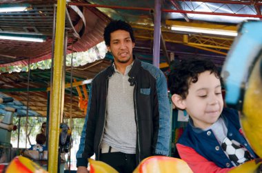 Afro Latin bekar baba, ayakta durup oğlunun elektrikli solucana binip parkta mekanik oyunlar oynamasını izlerken mutlu ve düşünceli.