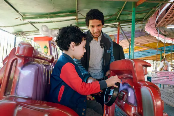 young afro latin venezuelan father standing telling his son how to play in mechanical car in electric games inside park, family concept, copy space