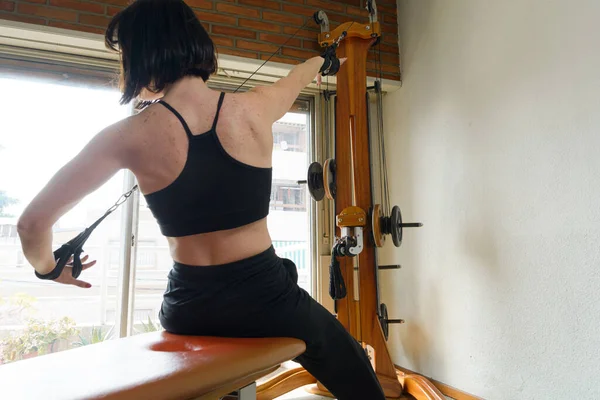Unrecognizable adult caucasian woman in black sportswear sitting training shoulders, back and arms with healthy exercise machine at home, training to compete and have healthy body.