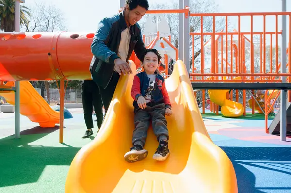 young latin man happy single father playing with his young son on slide in public park at noon, sharing with family on weekend