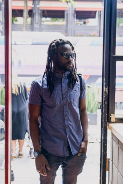 Afrika kökenli erkek müşterinin dikey görüntüsü. Rastalı, güneş gözlüklü, günlük kıyafetleri olan, ayakta durup kafeye giren...