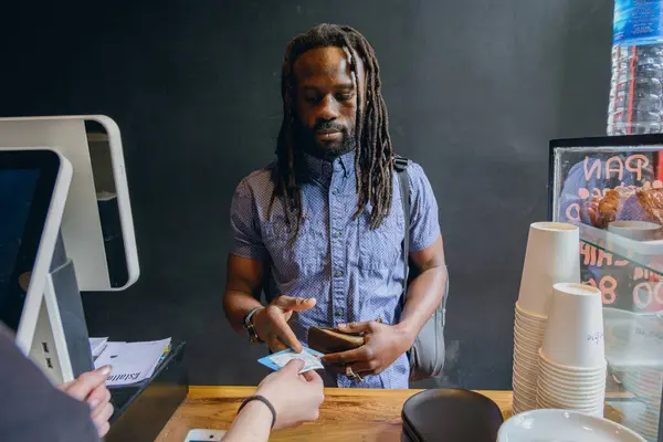 young black man with dreadlocks and beard standing in front of restaurant counter receiving his cards from waiter after paying for food order