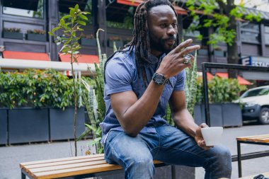 Saçlı, sakallı ve gömlekli genç afro adam dışarıda oturmuş biriyle konuşuyor ve tartışıyor ve kahve içiyor.