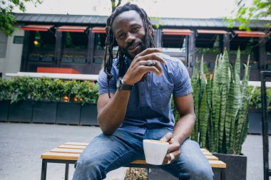 Sakallı ve rastalı genç Afrikalı adam mutlu gülümsüyor, konuşuyor ve kahve içiyor. Kafenin dışındaki ahşap bankta oturuyor, boşluğu kopyalıyor..