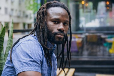Sakallı, rastalı ve gömlekli Afrikalı bir adamın profil portresi, kafenin dışında oturmuş yalnız ve sakin bir şekilde kameraya bakıyor, boşluğu kopyalıyor..