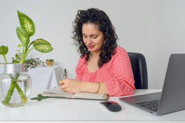Latin bir iş kadınının masasında oturup deftere yazarak yapması gereken işleri yazması ön planda..