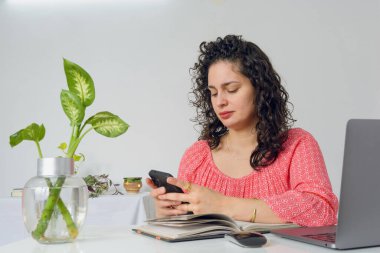 Bigudileri ve iş kıyafetleri olan genç Latin bir kadın ev ofisinde çalışıyor, telefonla mesajlar gönderiyor ve bildirileri ve sosyal ağları kontrol ediyor. Boşluğu kopyala.