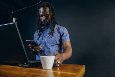 Rastalı ve sakallı Afrikalı siyahi bir adam, serbest meslek sahibi, kafede telefon kullanıyor, kahve içiyor, internet üzerinden müşterilerine hizmet ediyor..
