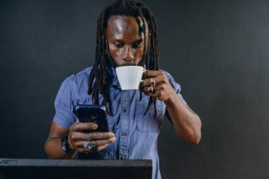 Genç Afrikalı iş adamı, rastalı ve sakallı, müşterilerine internet işlerinde hizmet vermekle meşgul, dizüstü bilgisayarıyla çalışıyor ve telefon izlerken kahve içiyor..