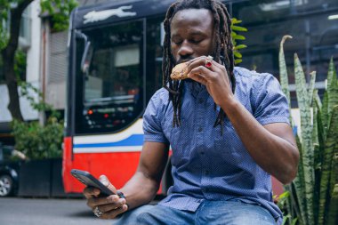 Rastalı ve sakallı genç siyahi Afrikalı adam cep telefonunu kullanarak oturuyor ve dışarıda kurabiye yiyor. yaşam biçimleri kavramı, kopyalama alanı