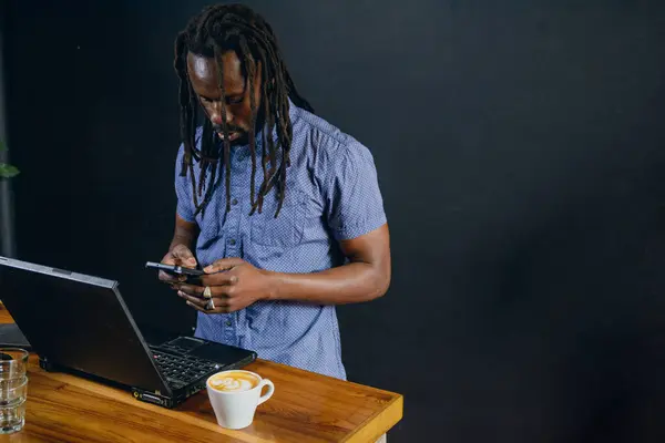 black business man with dreadlocks, beard and casual clothes, standing inside cafe busy working using phone sending messages, copy space