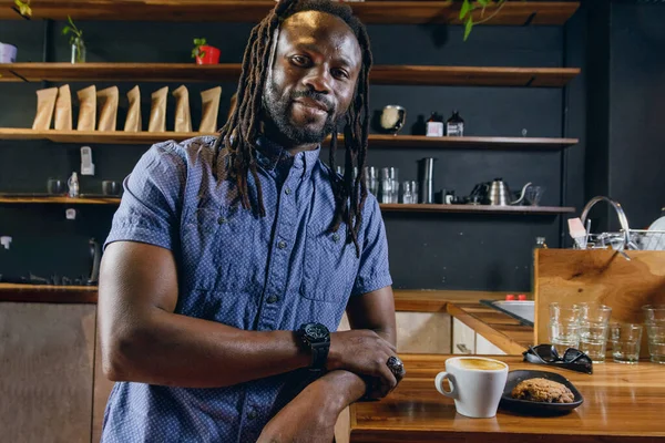 black man with dreadlocks and beard, eating coffee with cookies, happy smiling looking at camera standing next to counter inside restaurant, copy space