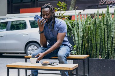 Rastalı Afrikalı bir adam, dışarıda oturmaktan mutlu, telefonundan sesli mesaj gönderiyor, dinlenirken ve kahve içerken kız arkadaşıyla iletişim kuruyor. Boşluğu kopyala.