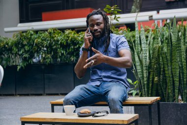 Rastalı ve sakallı Afrikalı bir adam, dışarıda oturuyor, telefon görüşmesi, teknoloji ve yaşam tarzı konsepti hakkında konuşuyor, kopyalama alanı..