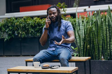 Rastalı ve sakallı Afrikalı bir adam, dışarıda oturuyor, telefon görüşmesi, teknoloji ve yaşam tarzı konsepti hakkında konuşuyor, kopyalama alanı..