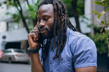 Rastalı ve sakallı Afrikalı bir adamın yakın çekimi, açık havada oturup, telefon görüşmesi, teknoloji ve yaşam tarzı konsepti hakkında konuşmak, kopyalama alanı..