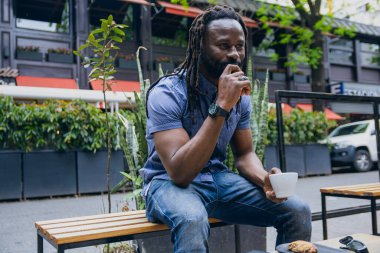 Genç Afrikalı bir adam dışarıda oturmuş kahve içiyor ve eli ağzında düşünüyor. İleriye bakıyor, meditasyon yapıyor ve dinlenmekten zevk alıyor..