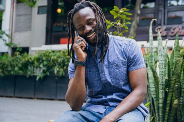 Güçlü ve yakışıklı Afrikalı bir adam, rastalı, sakallı ve tişörtlü, mutlu bir şekilde gülümseyen dışarıda oturmuş telefonla, hayat tarzıyla ve teknoloji konseptiyle konuşuyor..