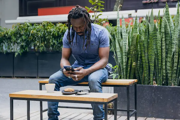 Stock image Strong young African man, wearing shirt and jeans and with dreadlocks, sitting outdoors texting on his phone, drinking coffee and eating cookies. copy space, lifestyles concept