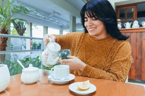young happy latin woman is alone sitting in restaurant with teapot pouring herself tea in porcelain cup during sunset, enjoying weekend