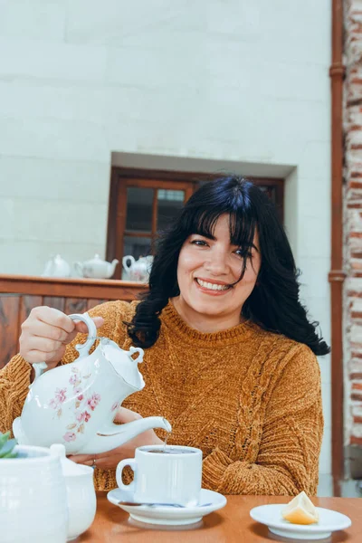 vertical image of happy and smiling young Latin woman, sitting in restaurant preparing her afternoon tea, resting and relaxed enjoying day