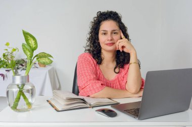 Ofisinde bukleli genç Latin bir kadının portresi. Kameraya bakıyor, mutlu, bir eli yüzüne yaslanmış, dizüstü bilgisayarı, telefonu ve defteri masanın üzerinde..