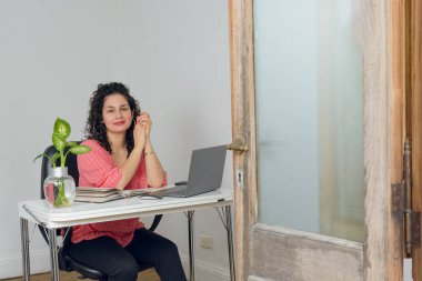 Bukleli genç, mutlu Latin kadın, yeni ofisinde oturuyor, gülümsüyor ve kameraya bakıyor, çok mutlu, girişimleri üzerinde çalışmaya hazır..