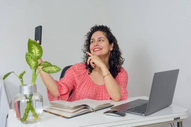 Venezuelalı kıvırcık saçlı genç bir Latin kadın ev telefonuyla telefonda gülümseyip bilgisayarını ve defterini masaya koymaktan mutlu. Boşluğu kopyala