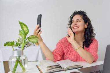 Venezuelalı kıvırcık saçlı genç bir Latin kadın ev telefonuyla telefonda gülümseyip bilgisayarını ve defterini masaya koymaktan mutlu. Boşluğu kopyala