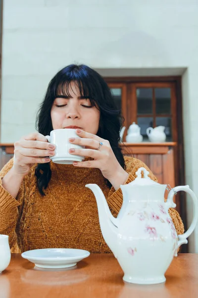 若いラテン語の女性の垂直画像 非常に暑いので 慎重に紅茶を飲んで座って 彼女の手で磁器のコップを保持しています — ストック写真