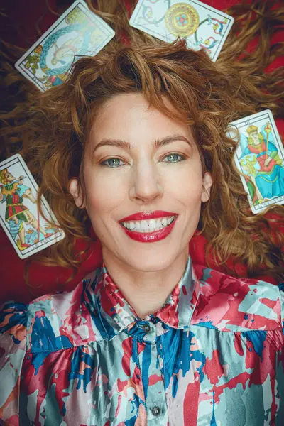 portrait of beautiful happy tarot reader woman smiling looking at camera lying on red table, with cards around her head and on her hair, vertical image