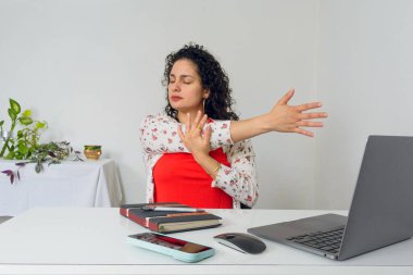 Genç Latin kadın ofisinde oturuyor, Gri laptopuyla işe gitmeden önce omuzlarını ve kollarını açıyor, Kırmızı ve beyaz giyinmiş, arka planda süs bitkileriyle dolu bir masa var.